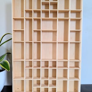 May include: A wooden display shelf with a grid of various sized compartments. The shelf is made of light-coloured wood and is designed to hold small items. The compartments are arranged in a rectangular pattern.