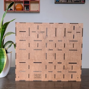 May include: A square, wooden panel with a geometric pattern of cut-out lines and rectangles. The panel is light brown with dark brown cut-outs. The words "SHELFISH MODERN" are printed in a rectangular box at the bottom. The panel is likely for home decor.