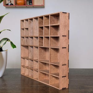 May include: A wooden display shelf with multiple square compartments. The shelf is made of light brown wood and has a grid-like structure. The edges of the shelf are reinforced with darker brown accents. The shelf is designed to hold small items.