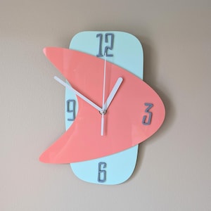 May include: A modern wall clock with a light blue and coral design. The clock face is divided into two sections, one with a light blue background and the other with a coral background. The numbers 12, 3, 6, and 9 are displayed on the clock face.