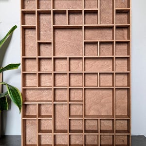 May include: A wooden display shelf for wall mounting, featuring a grid of compartments in various sizes. The light brown wood has a natural grain finish. The design incorporates a mix of square and rectangular spaces for display.