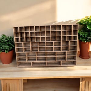 May include: A wooden display shelf with many small, square compartments. The shelf is made of light-coloured wood and sits on a wooden surface. Two potted plants are visible on either side of the shelf.
