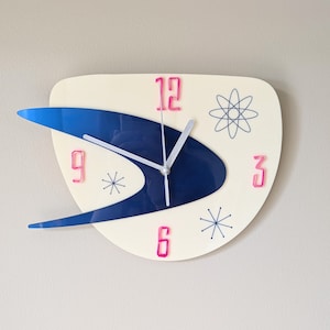 May include: A white and blue atomic-style clock with pink numbers. The clock has a blue and white design with a silver hand and a white background. The clock has a 12, 9, 3, and 6 on it.