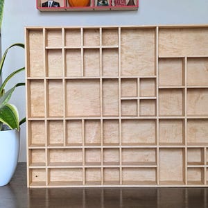 May include: A wooden display shelf with numerous compartments of varying sizes. The natural wood finish showcases the grain. The shelf is designed for organizing and displaying small items. It is a rectangular shape and sits on a dark surface.