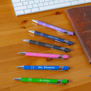 May include: A brown leather bound journal with a blue ribbon bookmark, a white keyboard, and six personalized pens with different colored barrels and silver accents. The pens are labeled with the names "Coach Rachel", "Joey & Pen", "Jane Doe Bakery", "Mr. Thomas", "Dr. Riche", and "Keep It Real".