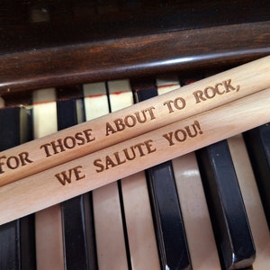 Puede incluir: Dos baquetas de madera con el texto "For those about to rock, we salute you!" grabado en ellas. Las baquetas est&aacute;n sobre un teclado de piano.