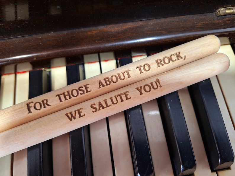 Puede incluir: Dos baquetas de madera con el texto "For those about to rock, we salute you!" grabado en ellas. Las baquetas est&aacute;n sobre un teclado de piano.