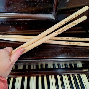 Puede incluir: Tres baquetas de madera con mensajes personalizados. La baqueta superior dice "To the best drummer I know! I love you Everlong, Rays." La baqueta del medio dice "FOR THOSE ABOUT TO ROCK, WE SALUTE YOU!"