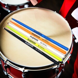 May include: A set of five drumsticks in various colors: black, lime green, white, orange, and blue. Each stick has the text "QuigBeats" and "5A Handpicked Premium Hickory" printed on it. The drumsticks are resting on a drum.