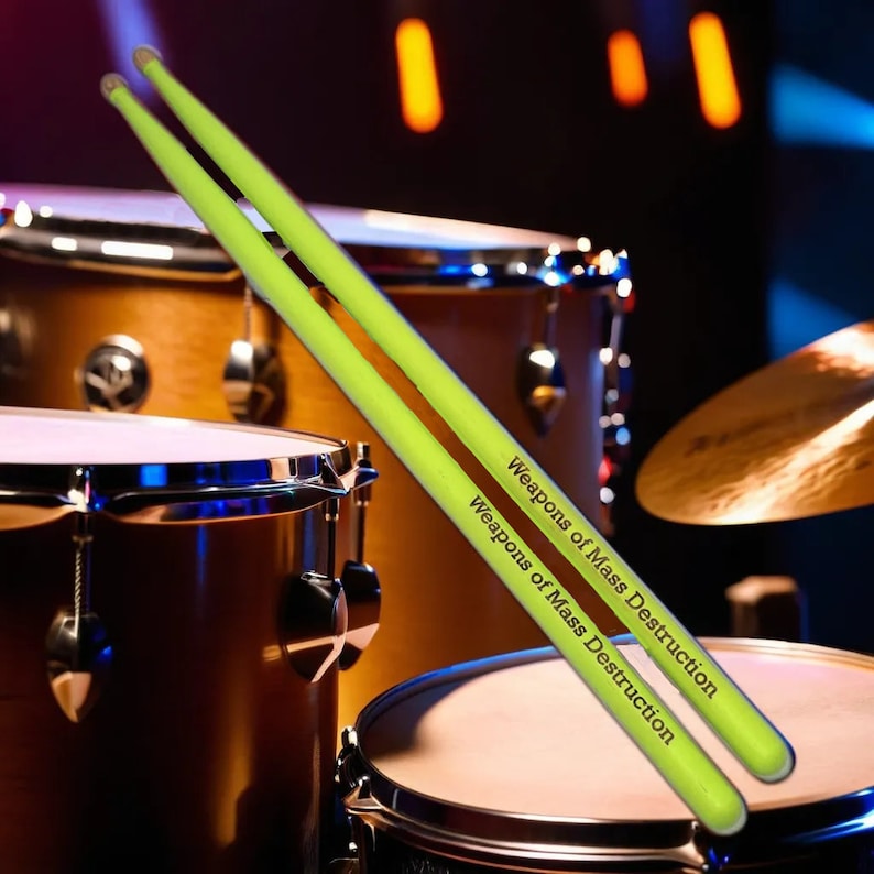 May include: Two neon green drumsticks with the text "Weapons of Mass Destruction" printed on them, resting on a drum set. The drumsticks are angled across the drum set, with the drums in the background.