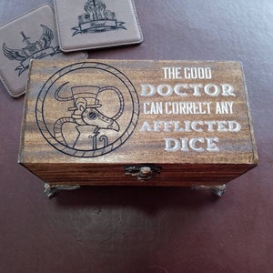 May include: A wooden box with a hinged lid and metal feet. The lid features a steampunk-style illustration of a doctor with a gas mask and the text "The Good Doctor Can Correct Any Afflicted Dice".