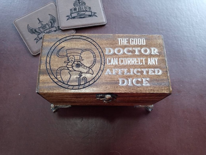 May include: A wooden box with a hinged lid and metal feet. The lid features a steampunk-style illustration of a doctor with a gas mask and the text "The Good Doctor Can Correct Any Afflicted Dice".