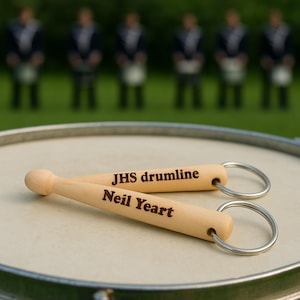 May include: Two wooden drumstick keychains with silver key rings rest on a drum. The text "JHS drumline" and "Neil Yeart" are engraved on the drumsticks. The background shows a group of people in uniform.