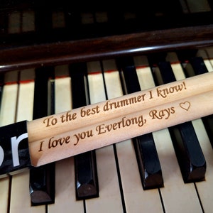 Puede incluir: Dos baquetas de madera con un mensaje personalizado: "To the best drummer I know! I love you Everlong, Rays" con un s&iacute;mbolo de coraz&oacute;n.