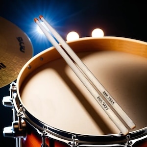 May include: A pair of white drumsticks with orange tips, each printed with the text "Tom's Sucks", are positioned over a drum. The drum has a light brown wooden frame and a tan drumhead. A cymbal is visible in the background.