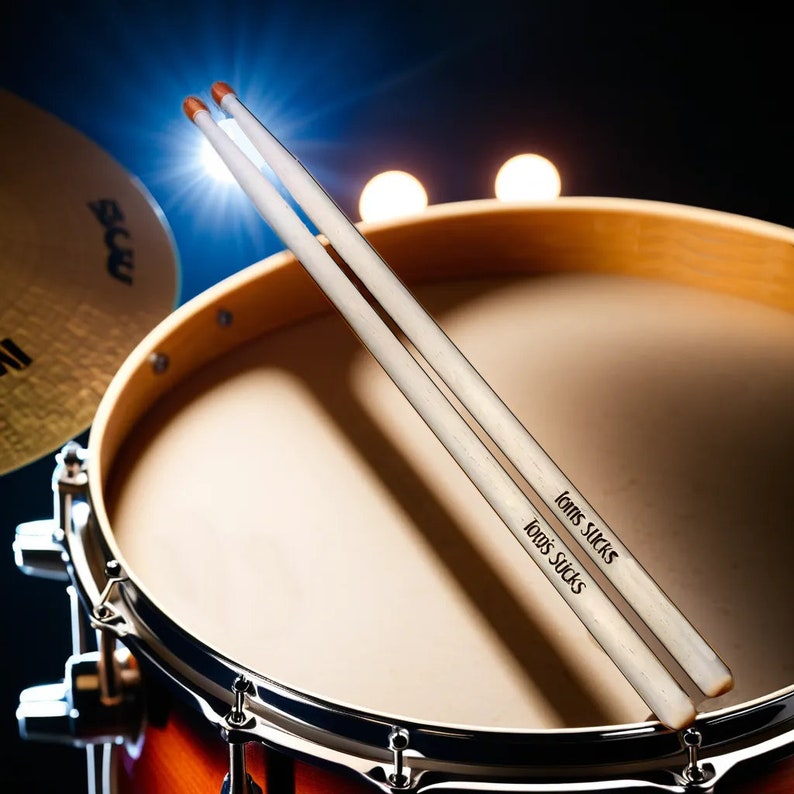 May include: A pair of white drumsticks with orange tips, each printed with the text "Tom's Sucks", are positioned over a drum. The drum has a light brown wooden frame and a tan drumhead. A cymbal is visible in the background.