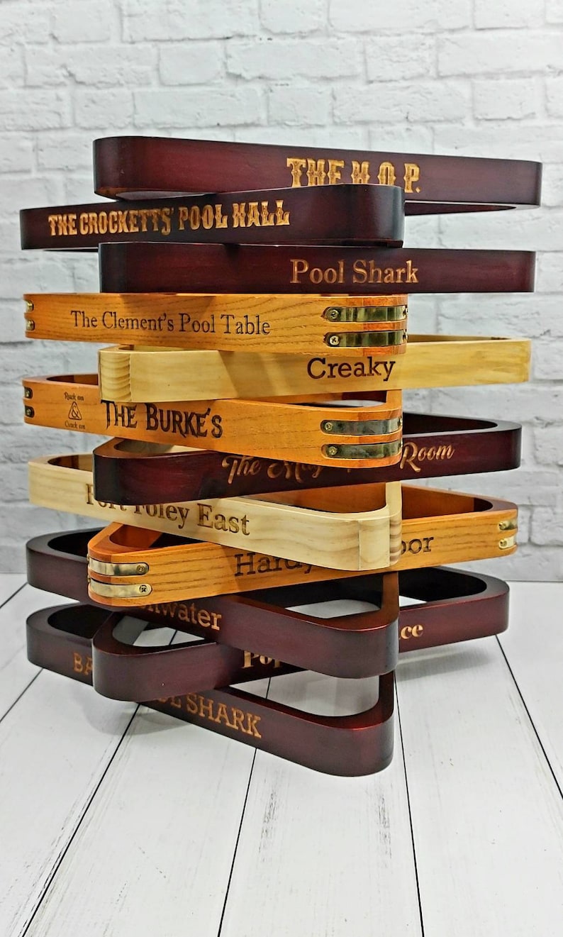 There is a stack of twelve pool triangles against a white background. There is a various amount of light and dark triangles and brass accented triangles. Each triangle is engraved with a different saying such as The Crocketts Pool Hall