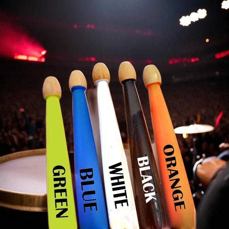 May include: A collection of five drumsticks in vibrant colors: green, blue, white, black, and orange. Each stick has a wooden tip and a colored shaft with the color name printed on it. The background shows a blurred concert setting.