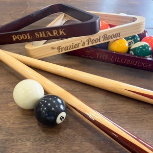 There are three pool triangles set on a table with pool balls and pool sticks laying around. One is engraved with Pool Shark one with The Frazier's Pool Room and one with a symbol that says rack em and crack em and The Lillibridges