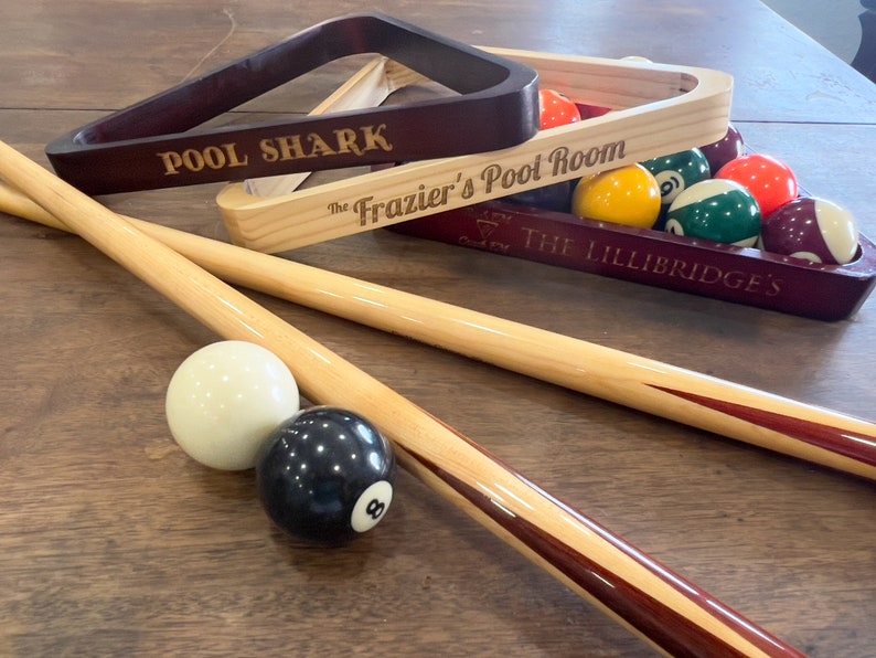 There are three pool triangles set on a table with pool balls and pool sticks laying around. One is engraved with Pool Shark one with The Frazier's Pool Room and one with a symbol that says rack em and crack em and The Lillibridges