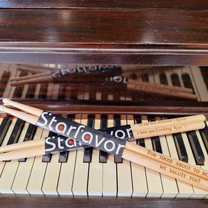 Puede incluir: Dos pares de baquetas de madera con mensajes personalizados. El par superior dice "To the best drummer I know! I love you Everlong, Rays." El par inferior dice "For those about to rock, we salute you!" Ambos pares est&aacute;n envueltos en un paquete negro con la palabra "Starfavor" impresa en &eacute;l.