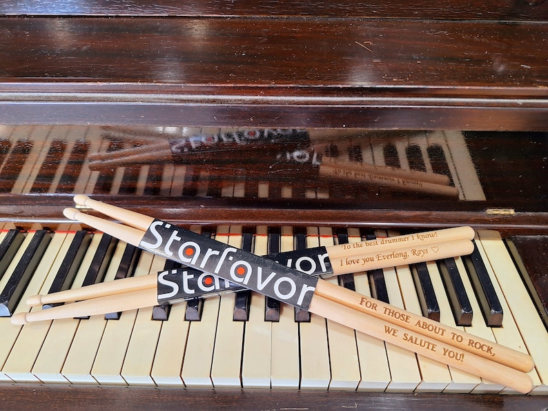 Puede incluir: Dos pares de baquetas de madera con mensajes personalizados. El par superior dice "To the best drummer I know! I love you Everlong, Rays." El par inferior dice "For those about to rock, we salute you!" Ambos pares est&aacute;n envueltos en un paquete negro con la palabra "Starfavor" impresa en &eacute;l.