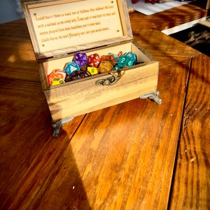 May include: A wooden box with a hinged lid, open to reveal a collection of colorful dice. The box has a brass latch and decorative feet. The inside of the lid has an inscription that reads: "Legend has it there's a magic box of holding that endows the user with a natural 20 on every roll. Long ago it was lost to time and serious players have been searching for it ever since. Could this be the box? Probably not, but you never know..."