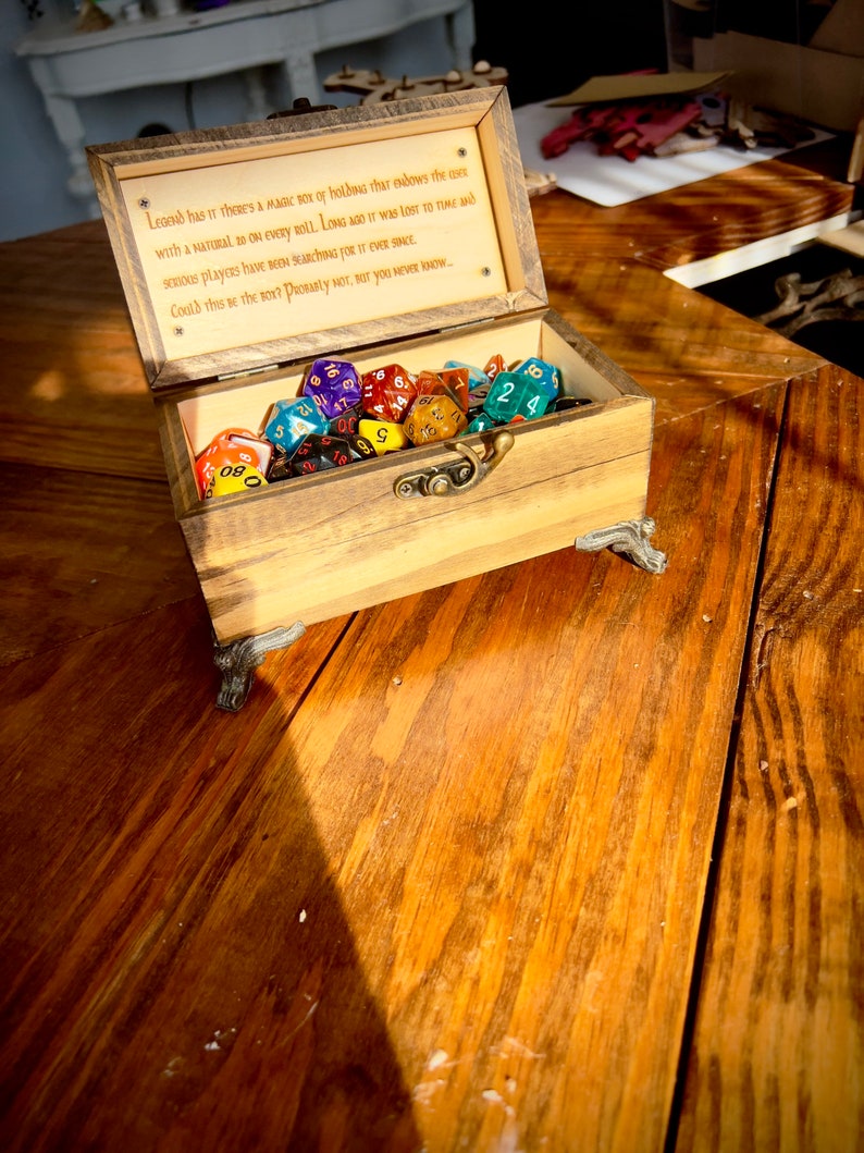 May include: A wooden box with a hinged lid, open to reveal a collection of colorful dice. The box has a brass latch and decorative feet. The inside of the lid has an inscription that reads: "Legend has it there's a magic box of holding that endows the user with a natural 20 on every roll. Long ago it was lost to time and serious players have been searching for it ever since. Could this be the box? Probably not, but you never know..."