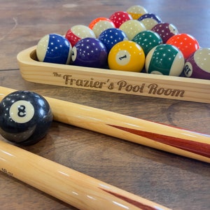 There is a light stained pool triangle set on a table filled with pool balls and set next to pool sticks. The pool triangle is laser engraved with The in small letters and Frazier's Pool Room in larger letters.