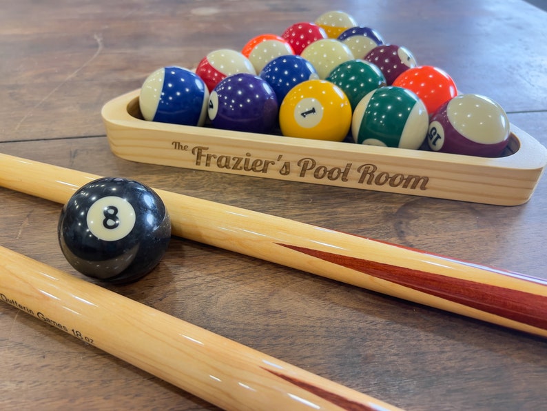 There is a light stained pool triangle set on a table filled with pool balls and set next to pool sticks. The pool triangle is laser engraved with The in small letters and Frazier's Pool Room in larger letters.
