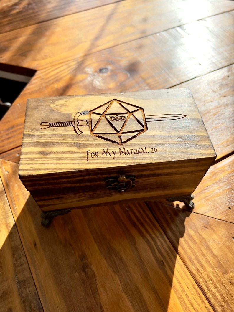 May include: A wooden box with a hinged lid and metal feet. The box is decorated with a laser-engraved image of a 20-sided die with a sword through it. The text "For My Natural 20" is also engraved on the box.