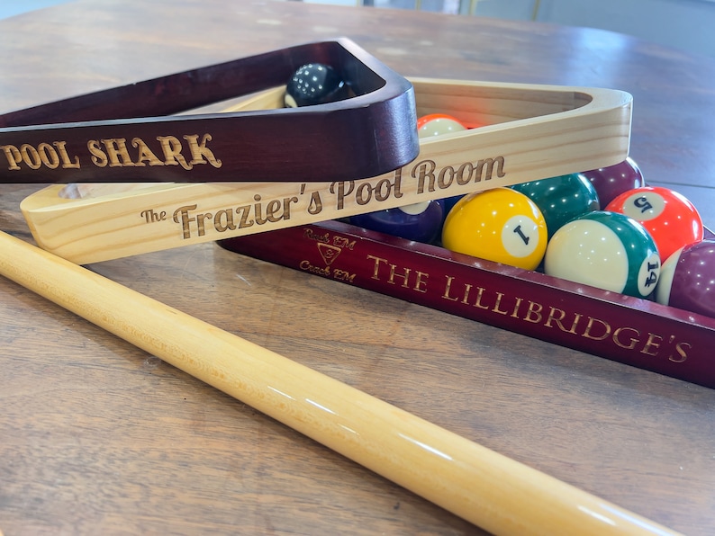 There are three pool triangles set on a table with pool balls and pool sticks laying around. One is engraved with Pool Shark one with The Fraziers Pool Room and one with a symbol that says rack em and crack em and The Lillibridges