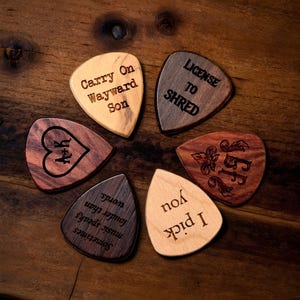 May include: A collection of six wooden guitar picks arranged in a circular pattern. Each pick features engraved text or designs, including phrases like "Carry On Wayward Son" and "License to Shred," and initials. The picks vary in wood tone.
