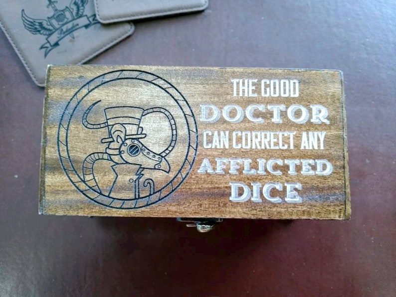 May include: Wooden box with a hinged lid. The box has a laser-etched design of a steampunk-style doctor with a top hat and goggles. The text on the box reads "The Good Doctor Can Correct Any Afflicted Dice".