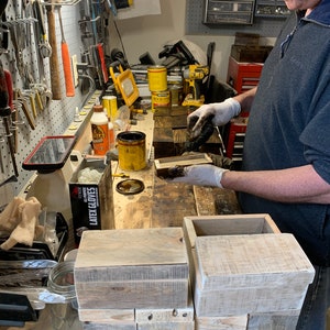 May include: A person wearing gloves applies a dark stain to a wooden box. The box is made of reclaimed wood and has a rustic look. There are several other wooden boxes stacked on a workbench in the background.