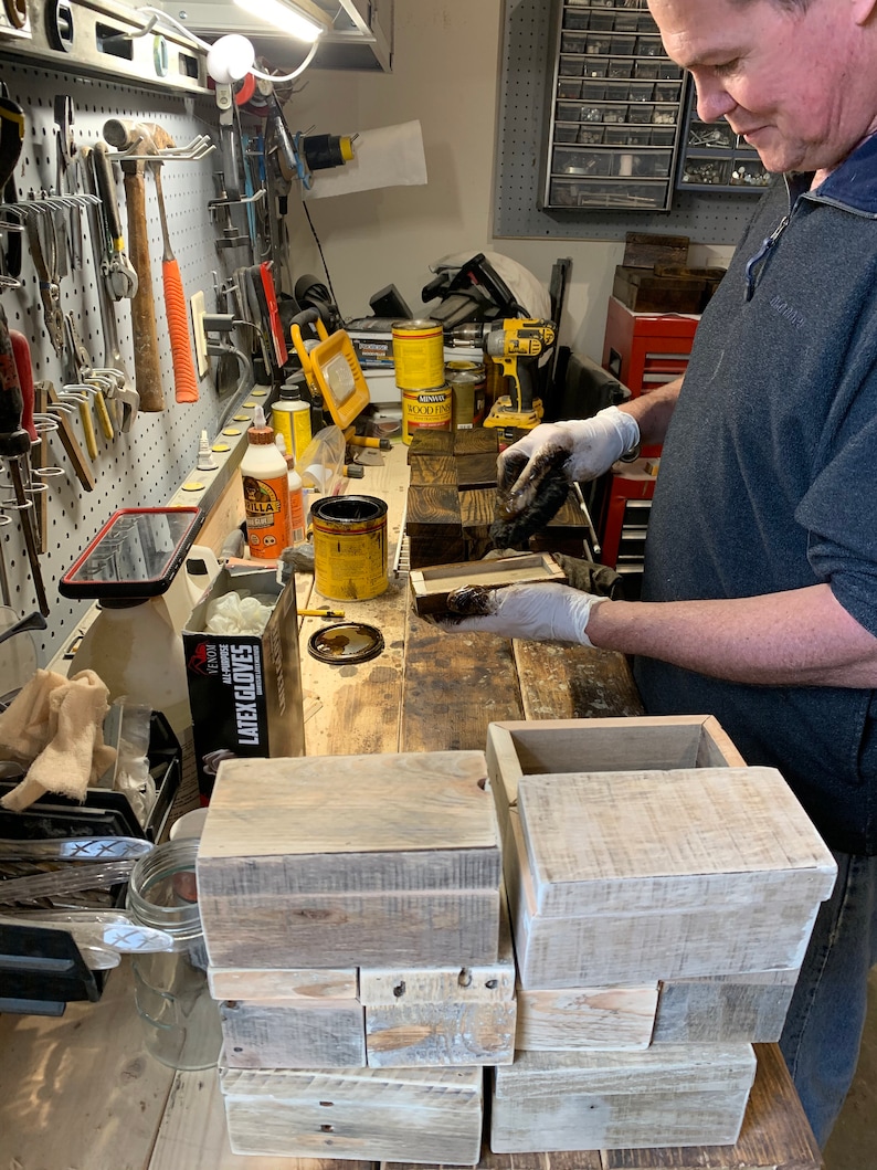 May include: A person wearing gloves applies a dark stain to a wooden box. The box is made of reclaimed wood and has a rustic look. There are several other wooden boxes stacked on a workbench in the background.