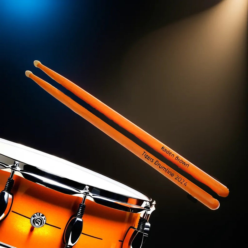 May include: Two orange drumsticks with white tips, angled towards the viewer, with the text "Kaden Brown Tigers Drumline 2024". A portion of an orange drum set is visible in the lower left corner against a dark background.