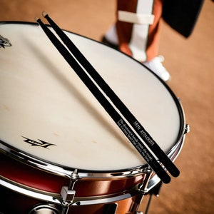 May include: A close-up of a drum with two black drumsticks resting on the drumhead. The drumsticks have text that reads "16TH ATTACK!!!" and "The drummer's hands create the music, but it's their heart that drives the beat."