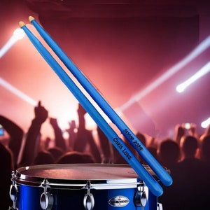 May include: Two blue drumsticks with the text "Senior 2024 Chris Long" are angled over a blue drum. The drumsticks have yellow tips. The drum is in front of a blurred background of a crowd and stage lights.
