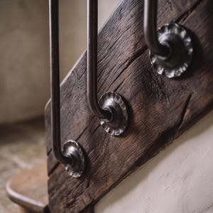 Può includere: Una trave di scala in legno scuro e usurato presenta tre supporti a parete circolari in metallo martellato. Ogni supporto fissa un montante di ringhiera in metallo scuro, mostrando una finitura rustica e testurizzata con venature del legno visibili.