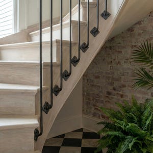 May include: A light-colored wooden staircase with black metal balusters and decorative accents. The stairs are set against a brick wall and a black and white checkered floor. A green plant is visible in the corner.