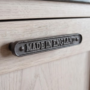 May include: A dark gray metal drawer pull with a textured background and raised lettering that reads MADE IN ENGLAND. The handle is horizontally mounted on a light-colored wood surface with a visible grain pattern and a horizontal seam.