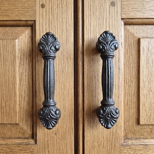 Puede incluir: Dos tiradores de armario de metal oscuro y texturizado con un centro estriado vertical y extremos decorativos en forma de abanico. Los tiradores están montados en una puerta de armario de madera de color marrón claro con veta visible.