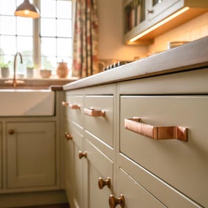 Può includere: Primo piano di un mobile da cucina con ante e cassetti verniciati di verde chiaro. I cassetti hanno maniglie color rame. La cucina ha un piano di lavoro in legno e una finestra sullo sfondo.