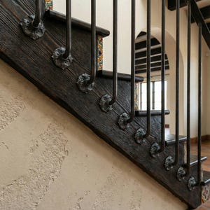 May include: A close-up of a wooden staircase with dark brown steps and decorative metal railing. The steps are adorned with ornate metal rosettes. The wall is a light beige color, and the staircase has a classic, rustic design.