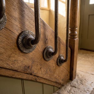 Può includere: Una scala caratterizzata da una trave in legno naturale con una venatura prominente. Fissati al legno, diversi supporti per balaustre in metallo scuro martellato presentano un design circolare testurizzato, ognuno dei quali sostiene un'asta verticale.