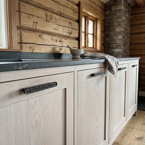 May include: Light-colored wooden kitchen cabinets featuring dark metal rectangular drawer pulls. Each pull is embossed with the text "MADE IN ENGLAND". A dark gray countertop sits above the cabinets, with a folded plaid cloth resting on top.