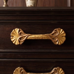 May include: A dark brown wooden dresser featuring two horizontal drawers. Each drawer is equipped with a decorative, textured gold-toned metal handle shaped like a stylized fan or shell with intricate carved details on the ends.