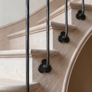 May include: Close-up of a wooden staircase with dark metal balusters. The steps are light-colored wood with a curved design. The balusters have a textured, hammered appearance and are attached with decorative, dark metal rosettes.