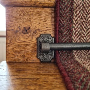 May include: A dark metal stair rod secured by an ornate, rectangular bracket with scrollwork detailing, mounted against a wooden staircase post. A section of a brown, green, and cream herringbone patterned stair runner carpet is visible.